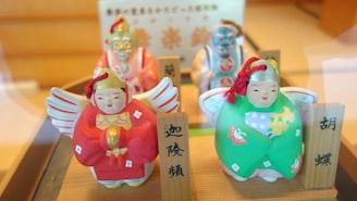 Four colorful ceramic figures with traditional attire are displayed, each with distinct features and vibrant colors. Two of the figures are dressed in red and green, with elaborate facial expressions and decorative patterns. The figures are accompanied by wooden name plaques with inscriptions in a non-Latin script.