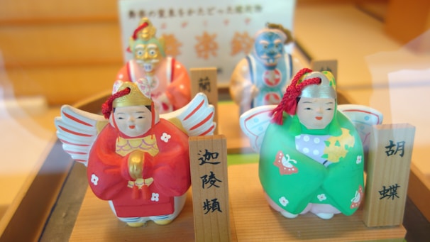 Four colorful ceramic figures with traditional attire are displayed, each with distinct features and vibrant colors. Two of the figures are dressed in red and green, with elaborate facial expressions and decorative patterns. The figures are accompanied by wooden name plaques with inscriptions in a non-Latin script.