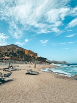 A sandy beach with scattered lounge chairs under an expansive blue sky with wispy clouds. A large resort hotel with vibrant orange and brown tones is situated on the left side, bordered by green palm trees. Waves gently lap against the shore, and a few people are visible enjoying the coastal setting.