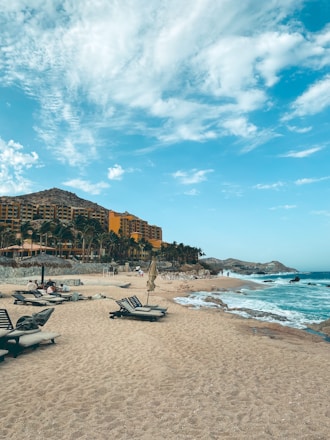A sandy beach with scattered lounge chairs under an expansive blue sky with wispy clouds. A large resort hotel with vibrant orange and brown tones is situated on the left side, bordered by green palm trees. Waves gently lap against the shore, and a few people are visible enjoying the coastal setting.