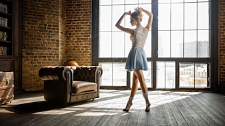 A dancer is poised elegantly in a well-lit room with large grid windows and a rustic vibe. The wooden floors and exposed brick walls add to the vintage atmosphere. An antique leather chair and a globe are visible in the background.