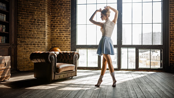 A dancer is poised elegantly in a well-lit room with large grid windows and a rustic vibe. The wooden floors and exposed brick walls add to the vintage atmosphere. An antique leather chair and a globe are visible in the background.