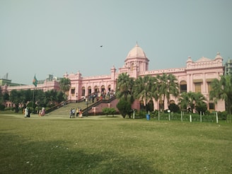 A large, historical building with a pink facade and a dome at the center is surrounded by palm trees and a manicured lawn. Several groups of people are visible near the entrance and around the building. A national flag is waving on a flagpole on the left side.