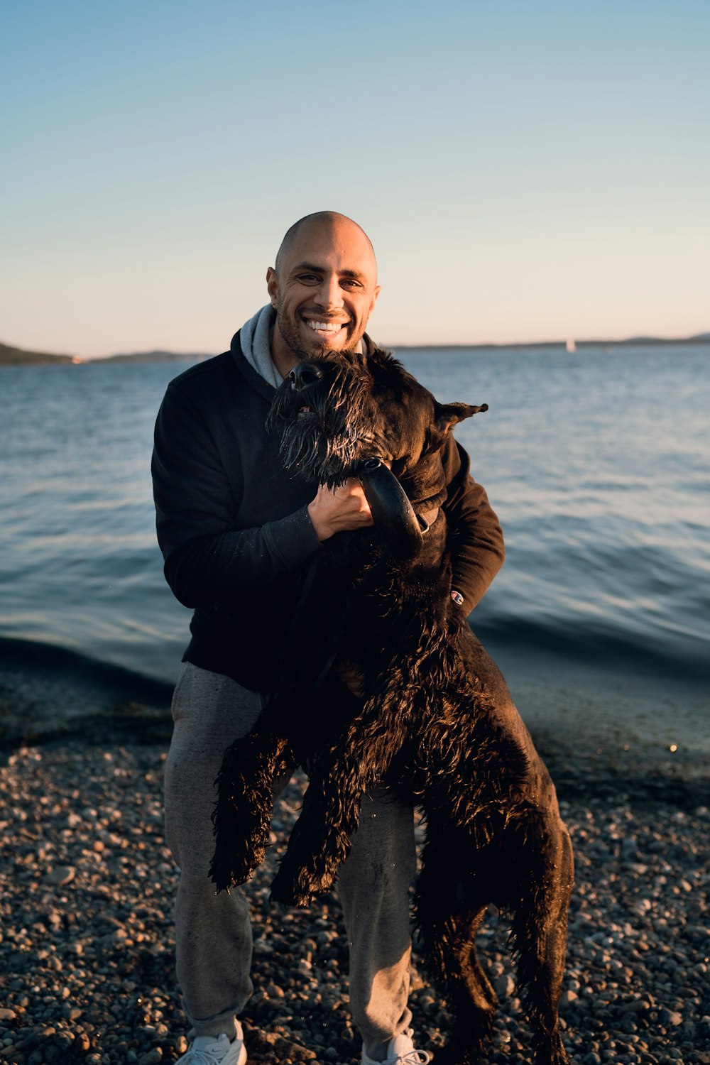 man in black jacket and blue denim jeans holding black dog on seashore during daytime