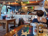 A live band is performing on a small stage in a rustic outdoor setting. The stage is adorned with string lights and a sign reading 'Shooters'. Patrons are seated at wooden tables, enjoying drinks like Lite and Corona, while a person in the foreground appears to be using a smartphone.