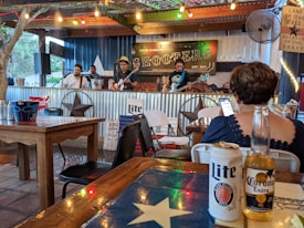 A live band is performing on a small stage in a rustic outdoor setting. The stage is adorned with string lights and a sign reading 'Shooters'. Patrons are seated at wooden tables, enjoying drinks like Lite and Corona, while a person in the foreground appears to be using a smartphone.