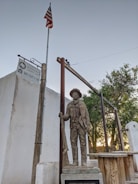 A statue of a historical figure stands near a wooden structure and a flagpole flying the American flag. Behind the statue, there is a sign indicating 'Old El Paso County Jail Museum.' The setting features some greenery, including a tree, and there are elements of rustic architecture.