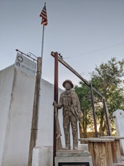 A statue of a historical figure stands near a wooden structure and a flagpole flying the American flag. Behind the statue, there is a sign indicating 'Old El Paso County Jail Museum.' The setting features some greenery, including a tree, and there are elements of rustic architecture.