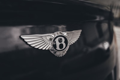 A close-up view of a luxury car emblem featuring a silver winged design with a central letter 'B'. The emblem is mounted on a glossy dark background, capturing the reflection of light.
