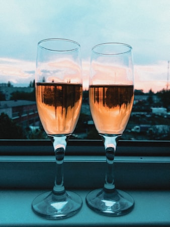 Two champagne glasses filled with a golden liquid are placed side by side on a window sill. The background reveals a scenic view of a sunset, casting warm hues across the sky.