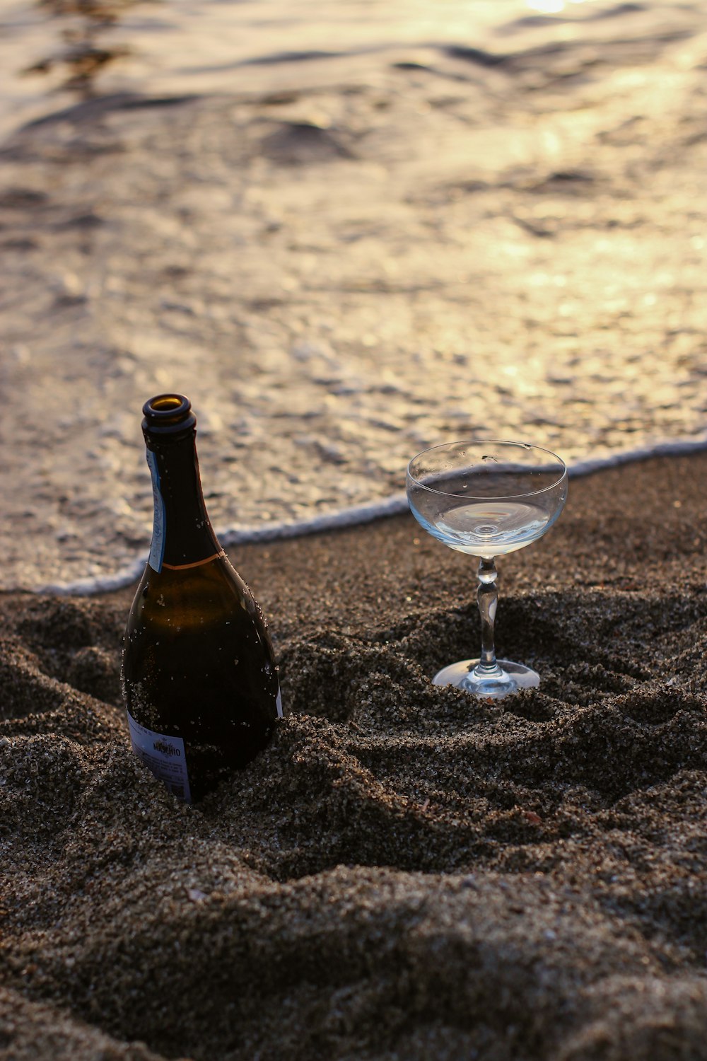 black glass bottle beside clear wine glass on brown sand during daytime