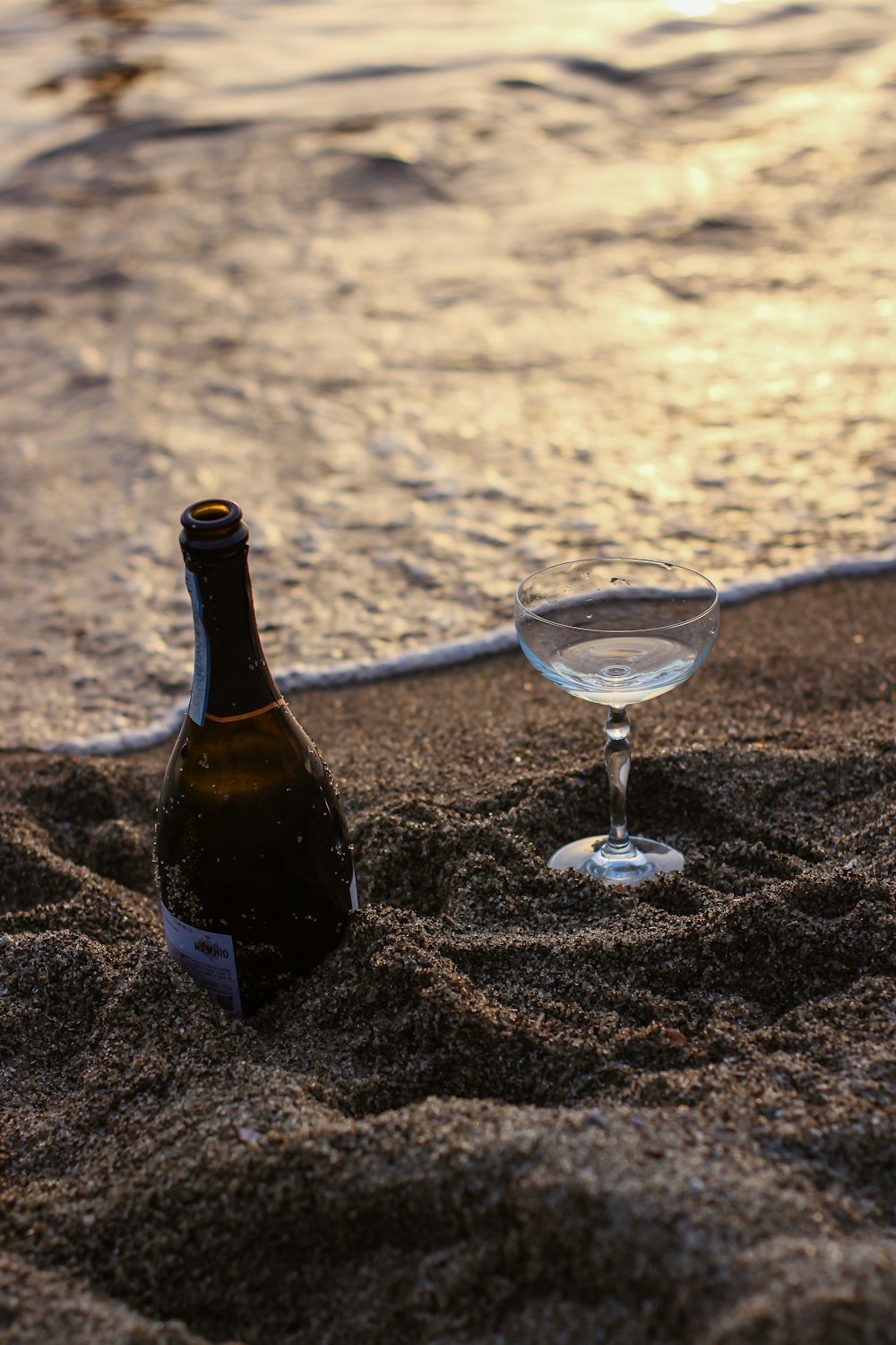 garrafa de vidro preto ao lado de vidro de vinho transparente na areia marrom durante o dia