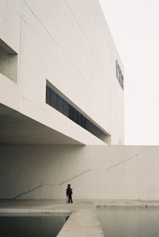 A minimalist architectural scene features a large, modern concrete building with geometric lines and an overhanging section. A narrow set of stairs ascends alongside the structure. Two figures stand on a wide path that juts out over a calm body of water, adding a human element to the stark landscape.