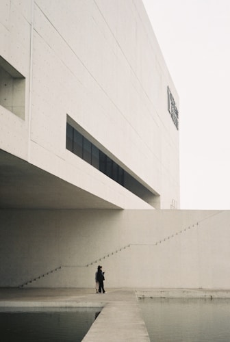 A minimalist architectural scene features a large, modern concrete building with geometric lines and an overhanging section. A narrow set of stairs ascends alongside the structure. Two figures stand on a wide path that juts out over a calm body of water, adding a human element to the stark landscape.