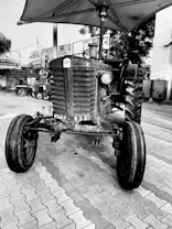 A black and white image of an old tractor parked on a paved surface. The tractor has prominent front wheels and a visible UTZ 93 license plate. An umbrella is attached above the tractor, providing shade. The background features various signs and a tree, along with some buildings and vehicles nearby.