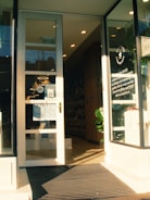 A storefront with large glass doors slightly open, leading into a well-lit interior. The entrance is marked with a sign displaying business details. Inside, shelves filled with products are visible, and a large plant is placed near the entrance. The lighting creates a warm, welcoming atmosphere.