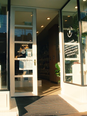 A storefront with large glass doors slightly open, leading into a well-lit interior. The entrance is marked with a sign displaying business details. Inside, shelves filled with products are visible, and a large plant is placed near the entrance. The lighting creates a warm, welcoming atmosphere.