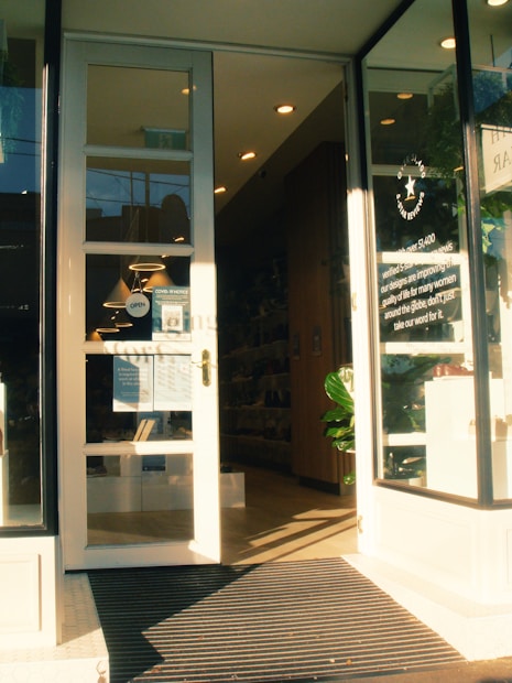 A storefront with large glass doors slightly open, leading into a well-lit interior. The entrance is marked with a sign displaying business details. Inside, shelves filled with products are visible, and a large plant is placed near the entrance. The lighting creates a warm, welcoming atmosphere.