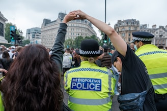 A police officer and a civilian are holding hands among a crowd in what appears to be a city setting. The officer is wearing a high-visibility jacket with 'Metropolitan Police' written on the back, and a hat. The surrounding crowd consists of people of various appearances, some wearing masks, and the background features urban architecture with multiple buildings.