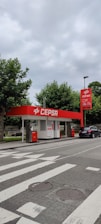 A small CEPSA gas station is situated by a roadside, featuring a distinct red and white color scheme. It includes a sheltered area with fuel pumps, and a car is parked nearby. Surrounding the station, there are lush green trees and a cloudy sky overhead.