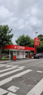 A small CEPSA gas station is situated by a roadside, featuring a distinct red and white color scheme. It includes a sheltered area with fuel pumps, and a car is parked nearby. Surrounding the station, there are lush green trees and a cloudy sky overhead.