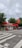 A small CEPSA gas station is situated by a roadside, featuring a distinct red and white color scheme. It includes a sheltered area with fuel pumps, and a car is parked nearby. Surrounding the station, there are lush green trees and a cloudy sky overhead.