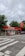 A small CEPSA gas station is situated by a roadside, featuring a distinct red and white color scheme. It includes a sheltered area with fuel pumps, and a car is parked nearby. Surrounding the station, there are lush green trees and a cloudy sky overhead.