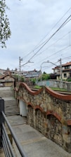 A stone footbridge with a red brick railing curves over a railway line, surrounded by residential buildings. Overhead power lines span across the train tracks. Trees and shrubs add greenery to the area, while stairs lead down to a paved path.