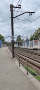 A train station platform with railway tracks running parallel to it. Overhead electrical wires are visible, supported by a tall metal post with a sign displaying the number 35 and the word 'ESPECIAL'. The platform is flanked by a blue and white building with red signage and surrounded by trees under a cloudy sky.