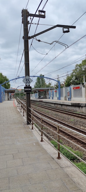 A train station platform with railway tracks running parallel to it. Overhead electrical wires are visible, supported by a tall metal post with a sign displaying the number 35 and the word 'ESPECIAL'. The platform is flanked by a blue and white building with red signage and surrounded by trees under a cloudy sky.