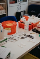 A table is cluttered with various medical supplies and equipment, including a red biohazard container, a spray bottle, packaged materials, and a tray with upright test tubes. The background features cabinets and chairs.