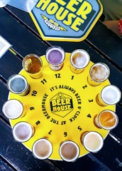 A circular yellow tray resembling a clock face features twelve glasses of beer, each placed at the positions of hourly markers. The tray is labeled 'BEER HOUSE' with the phrase 'It's always beer o'clock at the Beerhouse'. A placemat with the same theme is visible in the upper part of the image.