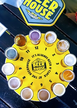 A circular yellow tray resembling a clock face features twelve glasses of beer, each placed at the positions of hourly markers. The tray is labeled 'BEER HOUSE' with the phrase 'It's always beer o'clock at the Beerhouse'. A placemat with the same theme is visible in the upper part of the image.