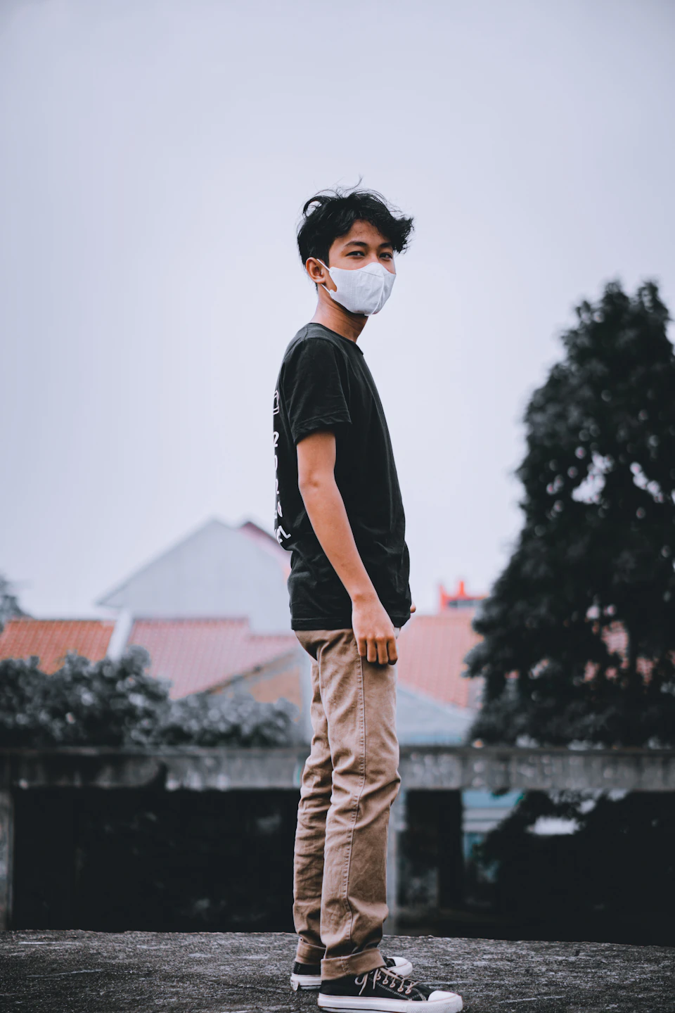 In the image, a young man is standing on a rooftop, wearing a black t-shirt and beige pants. He has his hands tucked into his pockets and appears to be looking off to the side. The rooftop he stands on is surrounded by trees, providing a natural backdrop to the scene.