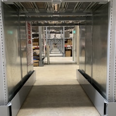 A long, narrow corridor with industrial metal shelving units on either side, extending into the distance with repetitive structural elements. The floor is made of polished concrete, and there are various boxes and items stored on the shelves. Overhead, there are metal racks and wire mesh, and some emergency exit signs with green lights are visible. The setting appears to be a warehouse or storage facility.