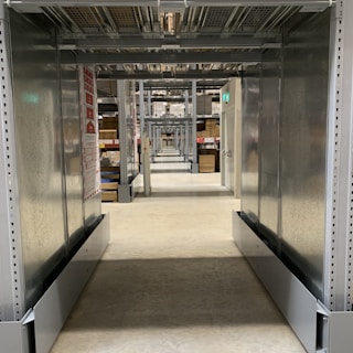 A long, narrow corridor with industrial metal shelving units on either side, extending into the distance with repetitive structural elements. The floor is made of polished concrete, and there are various boxes and items stored on the shelves. Overhead, there are metal racks and wire mesh, and some emergency exit signs with green lights are visible. The setting appears to be a warehouse or storage facility.