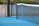 A close-up view of a blue hard surface tennis court featuring a black net stretched across it. The court is bordered by a metal fence with a concrete wall and vegetation in the background.