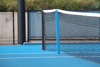 A close-up view of a blue hard surface tennis court featuring a black net stretched across it. The court is bordered by a metal fence with a concrete wall and vegetation in the background.