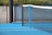 A close-up view of a blue hard surface tennis court featuring a black net stretched across it. The court is bordered by a metal fence with a concrete wall and vegetation in the background.