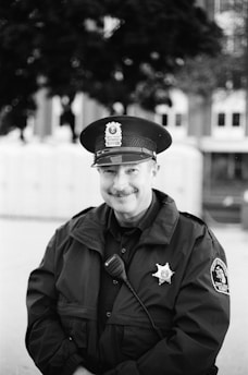 A uniformed police officer stands outdoors, wearing a badge and a cap with an emblem. The background is blurred, suggesting a setting in front of a building or public space.