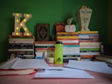 A study desk is arranged with a variety of books stacked in two piles. An illuminated letter 'K' sits on one pile. An assortment of colored books with Arabic titles is visible. A bright green water bottle with an Adidas logo is placed in front of the books. There are also papers, a pen, an orange clock, and a decorative lantern with tissue on the desk. The background wall is painted green.