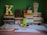 A study desk is arranged with a variety of books stacked in two piles. An illuminated letter 'K' sits on one pile. An assortment of colored books with Arabic titles is visible. A bright green water bottle with an Adidas logo is placed in front of the books. There are also papers, a pen, an orange clock, and a decorative lantern with tissue on the desk. The background wall is painted green.