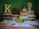 A study desk is arranged with a variety of books stacked in two piles. An illuminated letter 'K' sits on one pile. An assortment of colored books with Arabic titles is visible. A bright green water bottle with an Adidas logo is placed in front of the books. There are also papers, a pen, an orange clock, and a decorative lantern with tissue on the desk. The background wall is painted green.