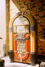 A vintage jukebox stands against a wall covered in brick-patterned wallpaper with green vine accents. The jukebox has a classic design with a rounded top, silver details, and vibrant colors, including red, yellow, and chrome.