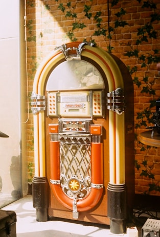 A vintage jukebox stands against a wall covered in brick-patterned wallpaper with green vine accents. The jukebox has a classic design with a rounded top, silver details, and vibrant colors, including red, yellow, and chrome.