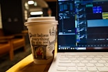 A paper coffee cup with an artistic design and the phrase 'Don't believe everything you hear' printed on it sits on a table next to an open laptop. The screen displays a video editing software with various tracks and timelines visible. The background features blurred indoor elements, creating a cozy atmosphere.
