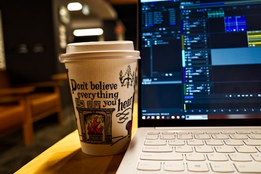 A paper coffee cup with an artistic design and the phrase 'Don't believe everything you hear' printed on it sits on a table next to an open laptop. The screen displays a video editing software with various tracks and timelines visible. The background features blurred indoor elements, creating a cozy atmosphere.