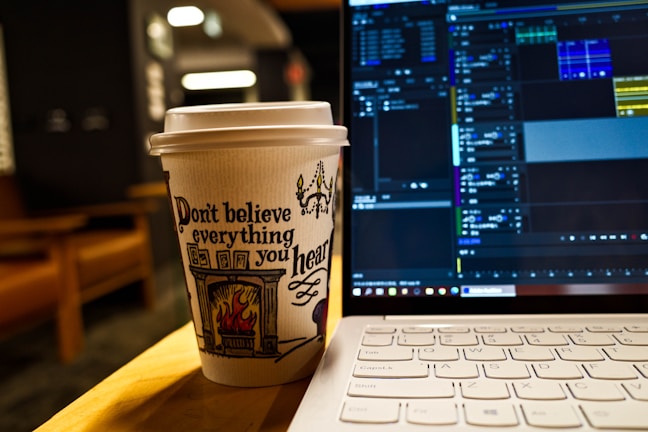 A paper coffee cup with an artistic design and the phrase 'Don't believe everything you hear' printed on it sits on a table next to an open laptop. The screen displays a video editing software with various tracks and timelines visible. The background features blurred indoor elements, creating a cozy atmosphere.