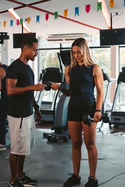 A woman is lifting dumbbells with guidance from a man in a gym setting. Various gym equipment such as treadmills are visible in the background along with colorful banners hanging from the ceiling.