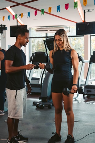 A woman is lifting dumbbells with guidance from a man in a gym setting. Various gym equipment such as treadmills are visible in the background along with colorful banners hanging from the ceiling.